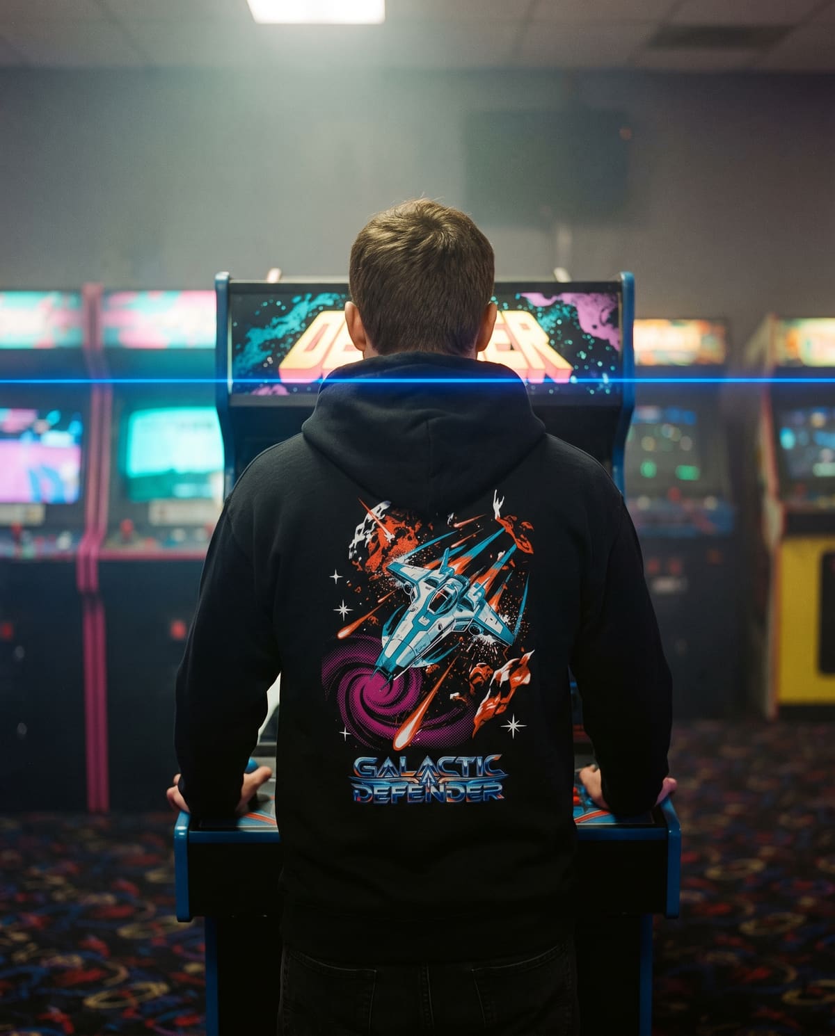 A man at a retro arcade cabinet at night, seen from behind in the black Interceptor hoodie with its spaceship back print