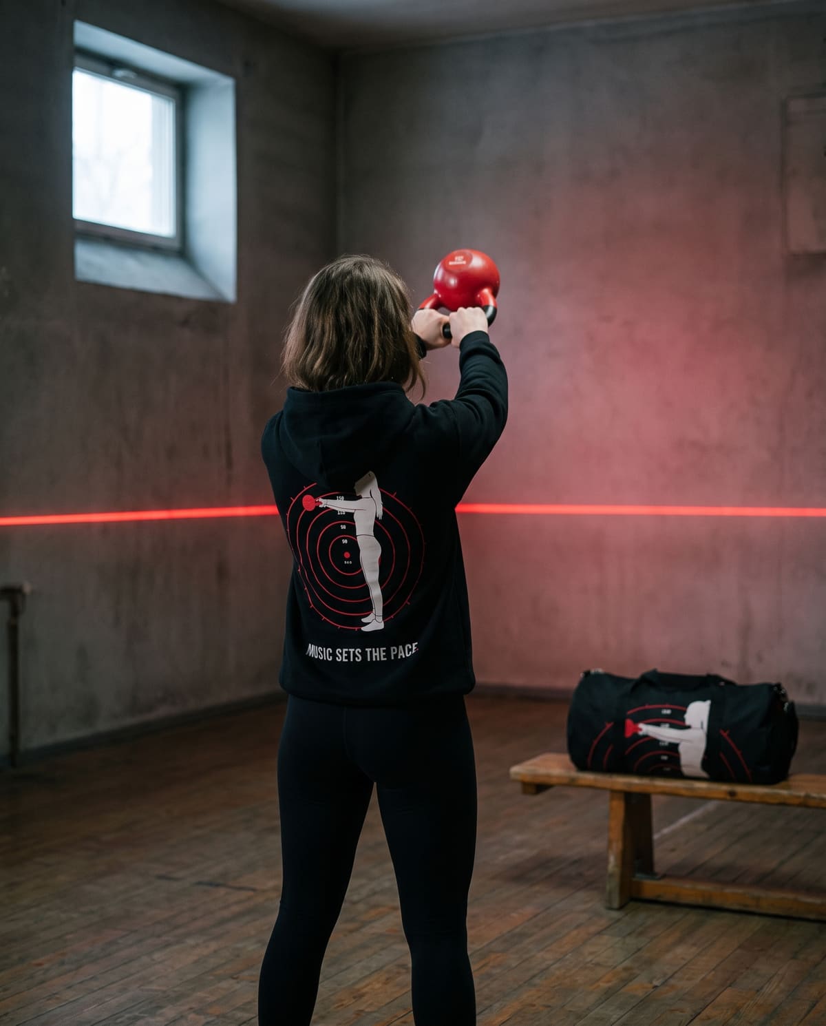 A woman mid kettlebell swing in a dark gym, seen from behind in the black Swing hoodie, the Gym Day duffel on the bench beside her